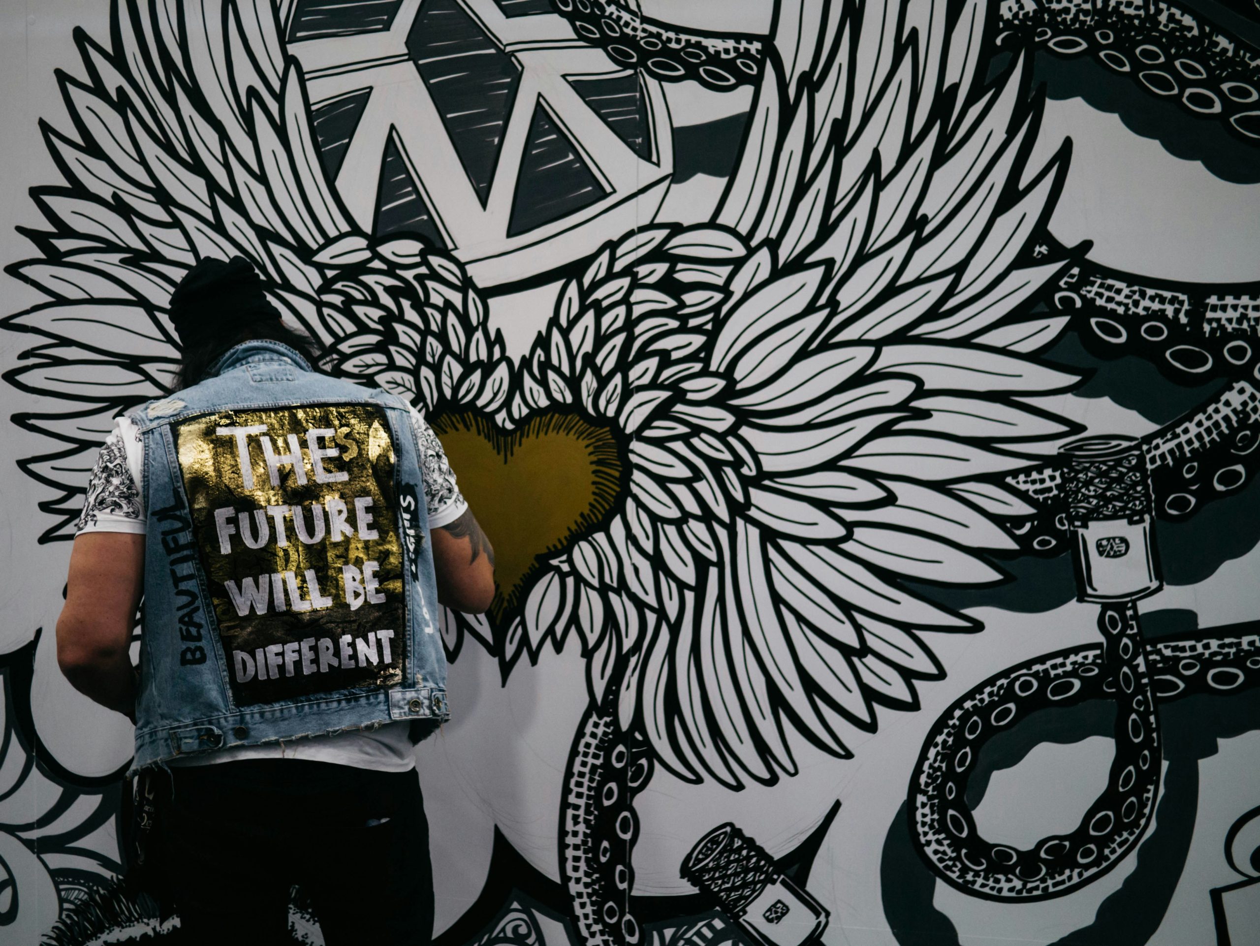 Man wearing a jacket that reads"THE FUTURE WILL BE DIFFERENT" painting a mural with a big, winged, gold heart.
